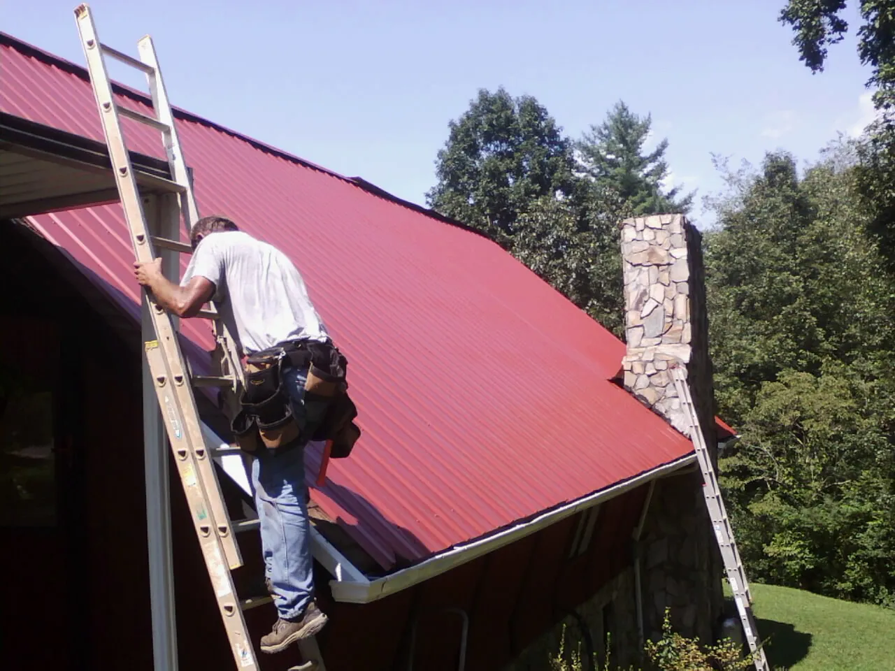Roofer on a ladder beside a red metal roof and stone chimney — Soffit & Fascia Repair by Roofing Brothers in Roswell